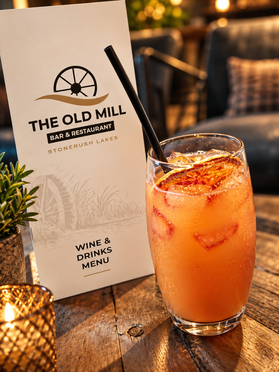 Peach-coloured cocktail in a tall curved glass with a dried orange garnish, beside The Old Mill drinks menu